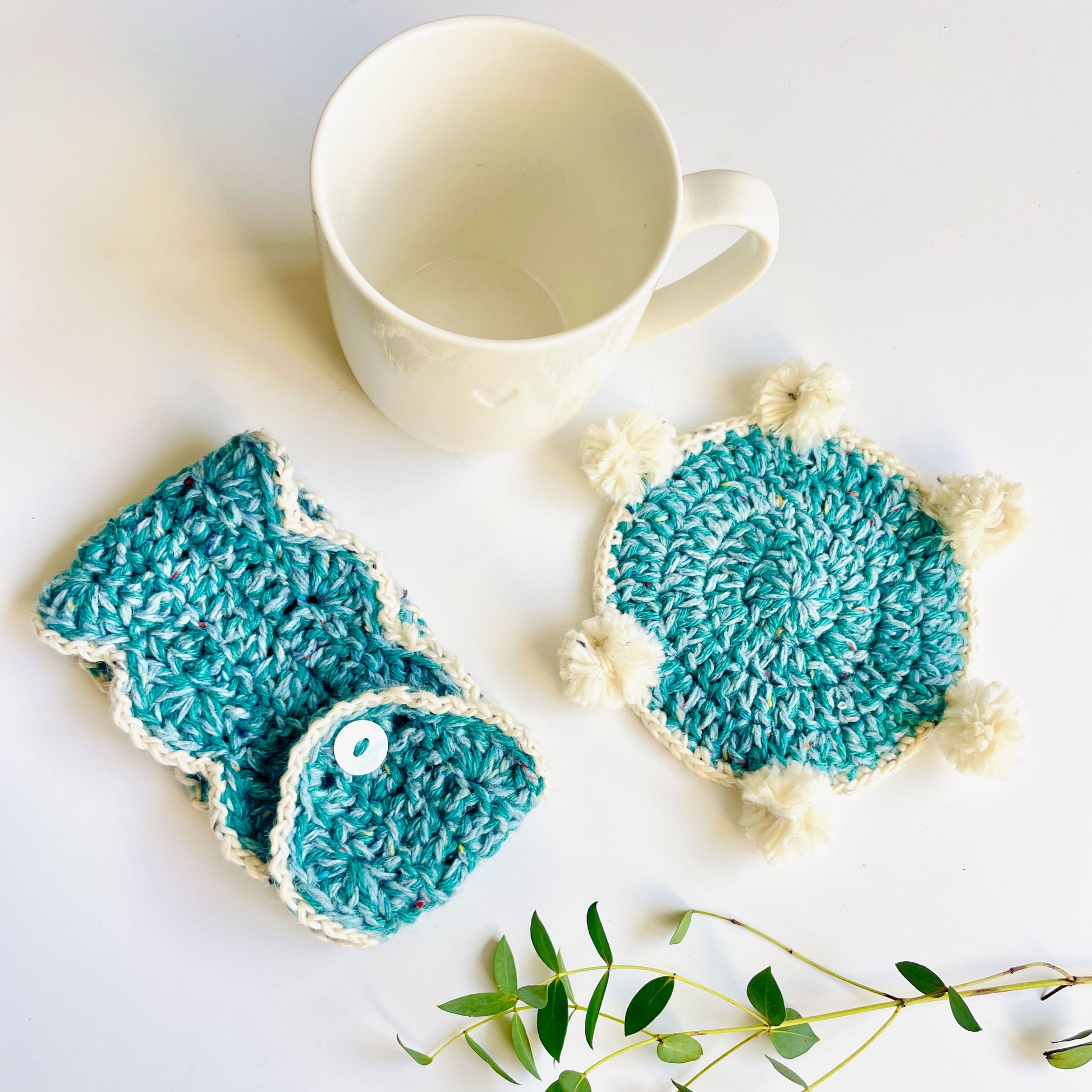 A crochet teal coaster and mug wrap with white trims on a white background with an off-white mug and foliage.