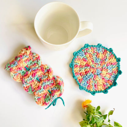 Colourful crochet coaster and mug wrap on a white surface with a mug and foliage.