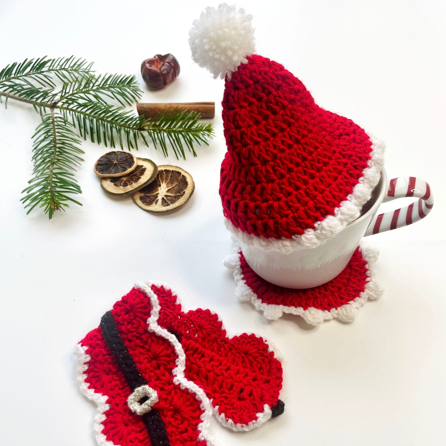 Matching crochet Santa coaster, mug wrap and hat set on a white surface with Christmas foliage.